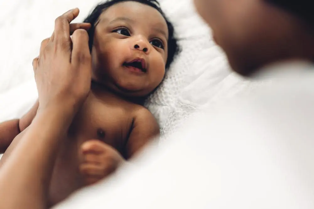 A baby lies on a white blanket, gazing up with a curious expression. A persons hand gently touches the babys cheek. The scene is soft and intimate, capturing a moment of tenderness.