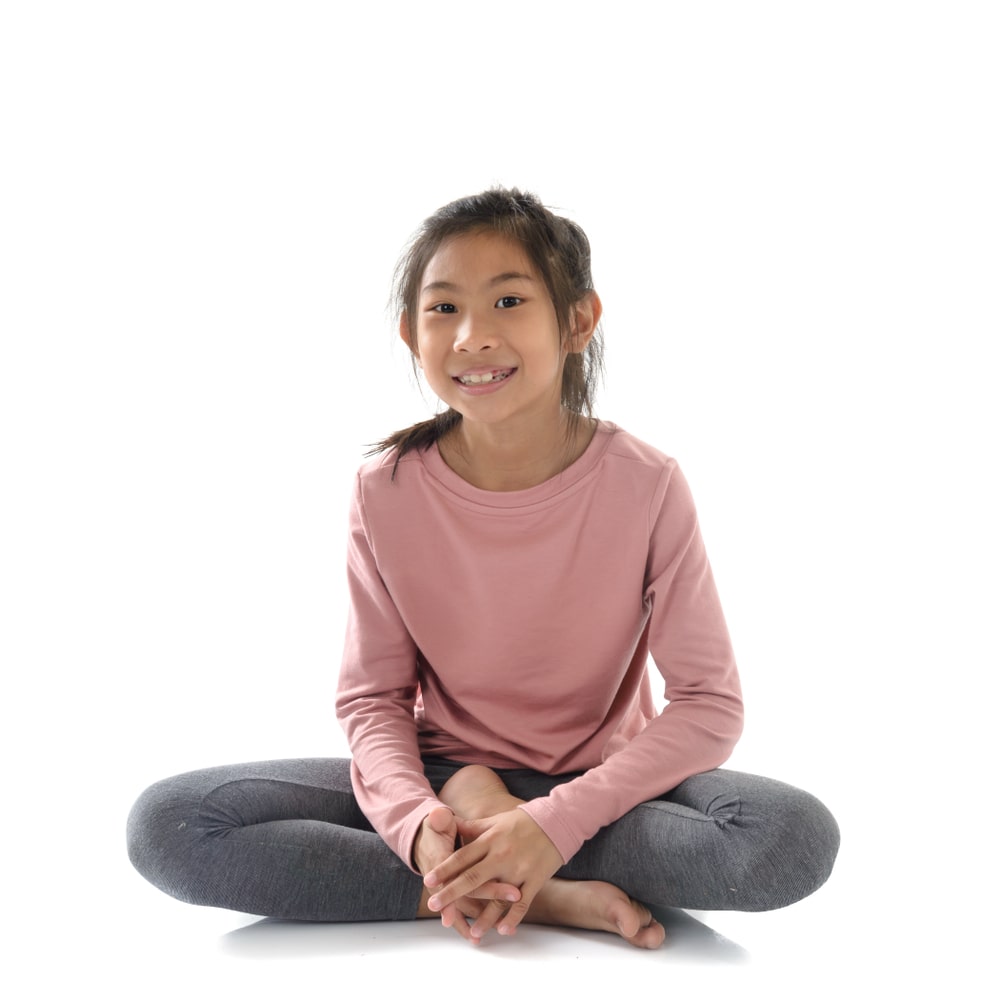 A young girl with dark hair is sitting cross-legged on the floor, smiling. She is wearing a pink long-sleeve top and gray pants. The background is white, creating a bright and simple setting.