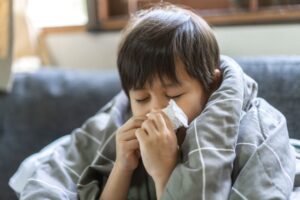 A young child with dark hair sits wrapped in a gray blanket, eyes closed, holding a tissue to their nose as if sneezing or blowing their nose, suggesting they are unwell.
