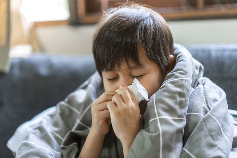 A young child with dark hair sits wrapped in a gray blanket, eyes closed, holding a tissue to their nose as if sneezing or blowing their nose, suggesting they are unwell.